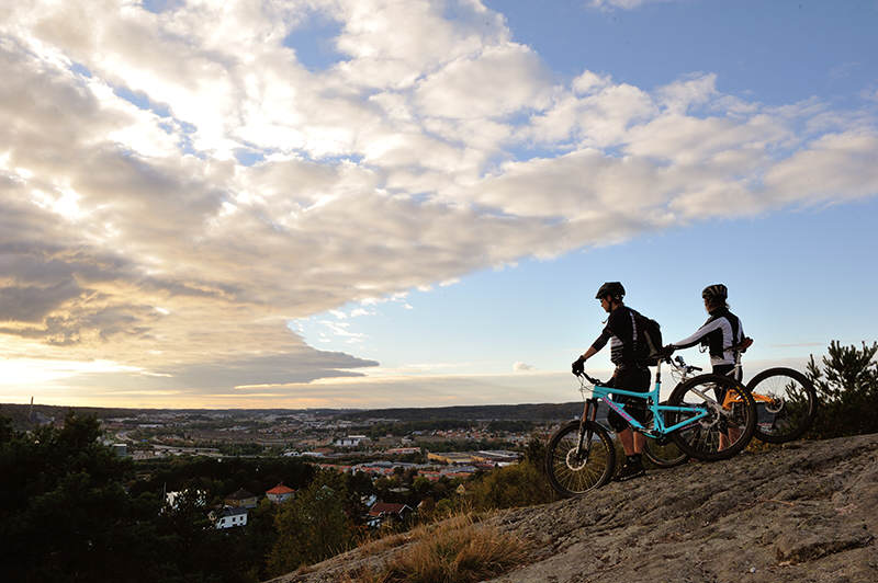 2 people with mountain bikes looking over houses as the sun goes down in a partially cloud filled sky