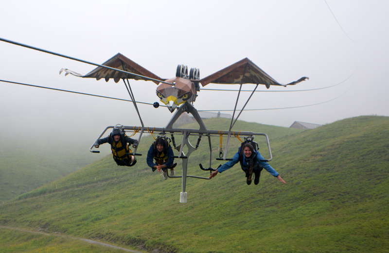 3 people on a zip line in the shape of a bird with grass hills behind and below