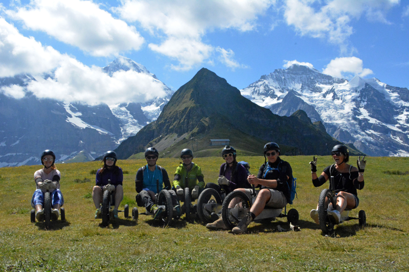 7 people sat on mountain carts on the grass in front of a pyramid shaped mountain with snow capped mountains behind