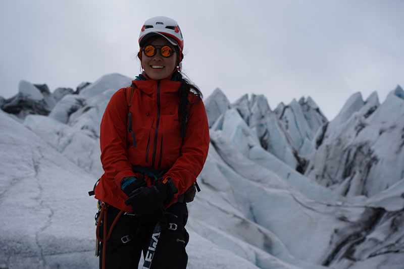 Female climber in red jacket, white helmet and Vallon Heron Glacier Glasses standing in front of ice pinnacles in Iceland