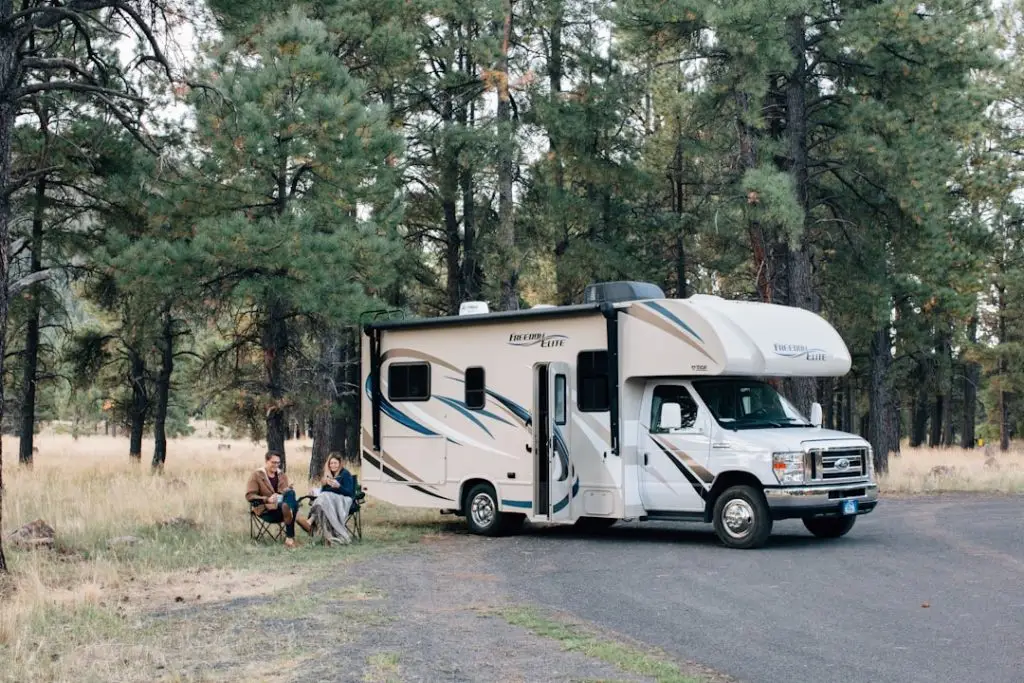 mobile home parked in woodland with a man and woman sitting near it in camping chairs